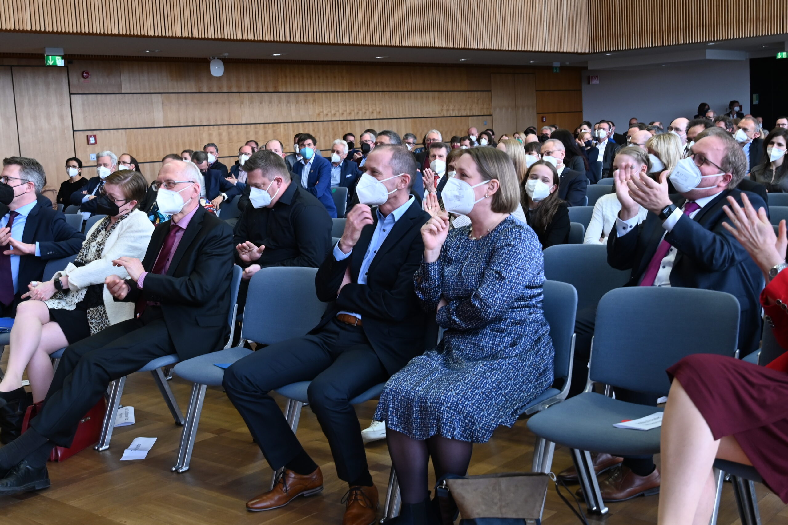 Eine Gruppe von Menschen in formeller Kleidung sitzt in Stuhlreihen in einem Konferenzraum der Hochschule Coburg. Die meisten tragen Gesichtsmasken und einige applaudieren. Der Hintergrund ist mit Holzvertäfelungen versehen, die der akademischen Versammlung einen Hauch von Eleganz verleihen.