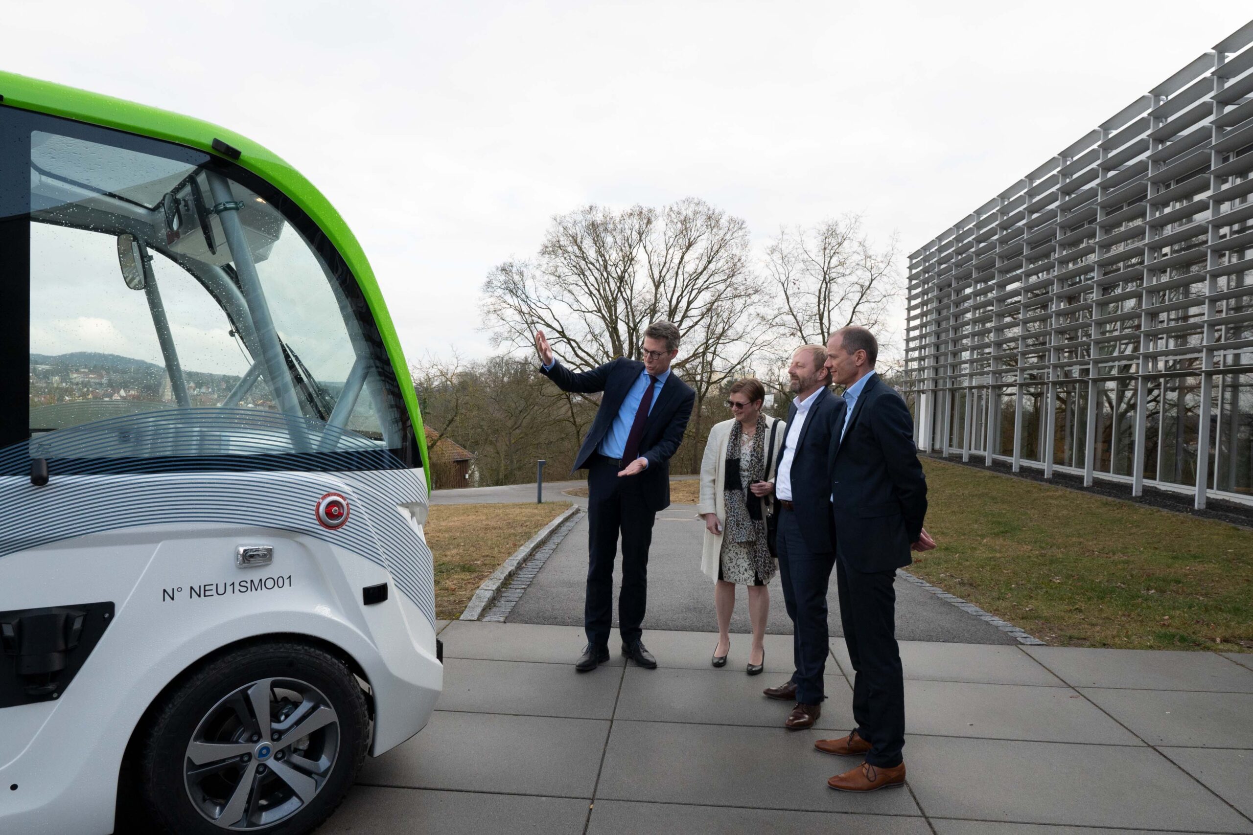 Vier Personen in Businesskleidung stehen vor der Hochschule Coburg und bewundern ein grün-weißes autonomes Fahrzeug. Ein Mann deutet auf das Fahrzeug, während die anderen aufmerksam zuhören. Im Hintergrund ergänzt ein modernes Gebäude die blattlosen Bäume.