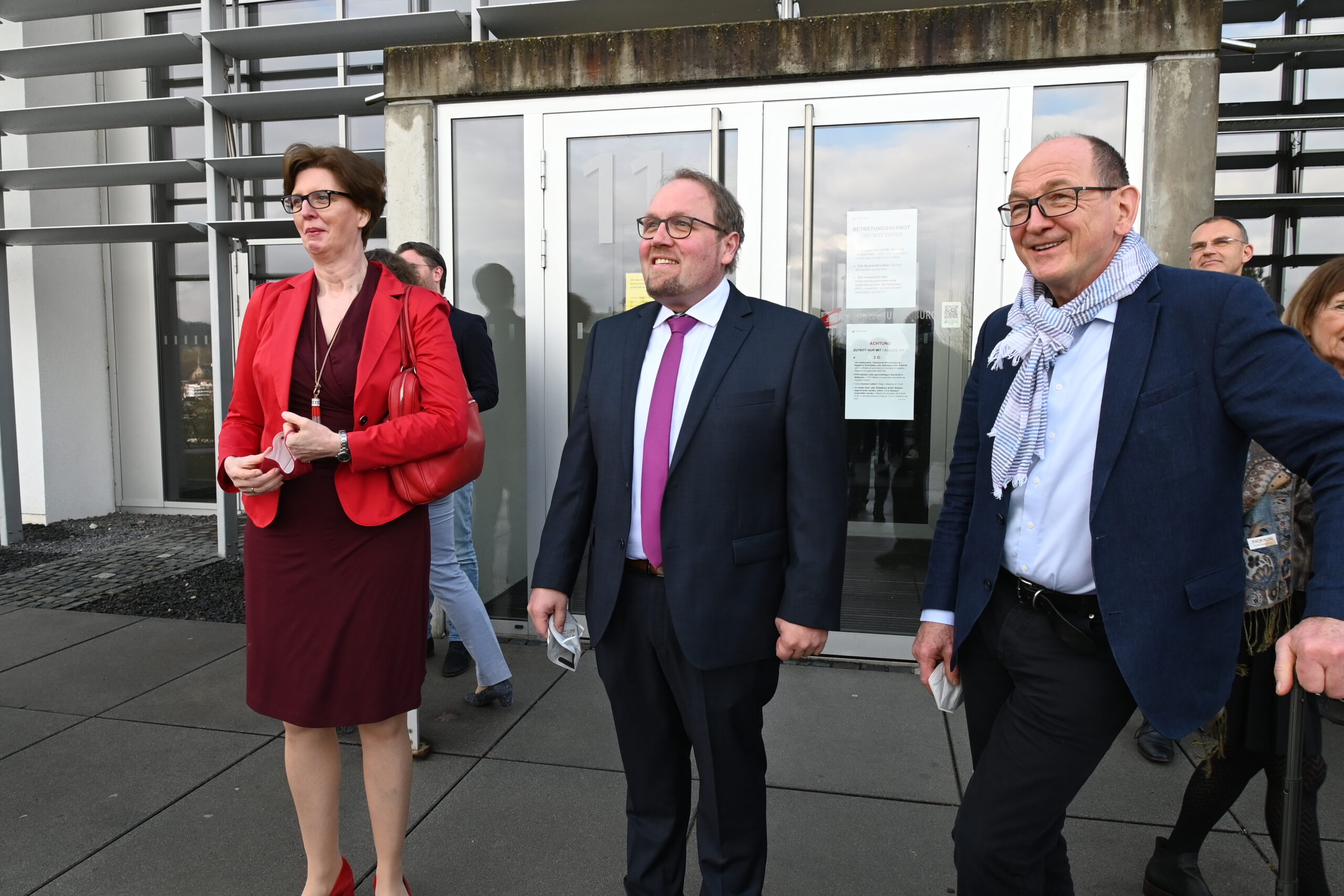 Drei Menschen stehen lächelnd vor einem Gebäude. Eine trägt ein rotes Jackett und einen roten Rock, eine andere einen Anzug mit rosa Krawatte und die dritte einen blauen Blazer mit Schal. Hinter ihnen hängen an der Glastür Aushänge mit Informationen zu Veranstaltungen der Hochschule Coburg.