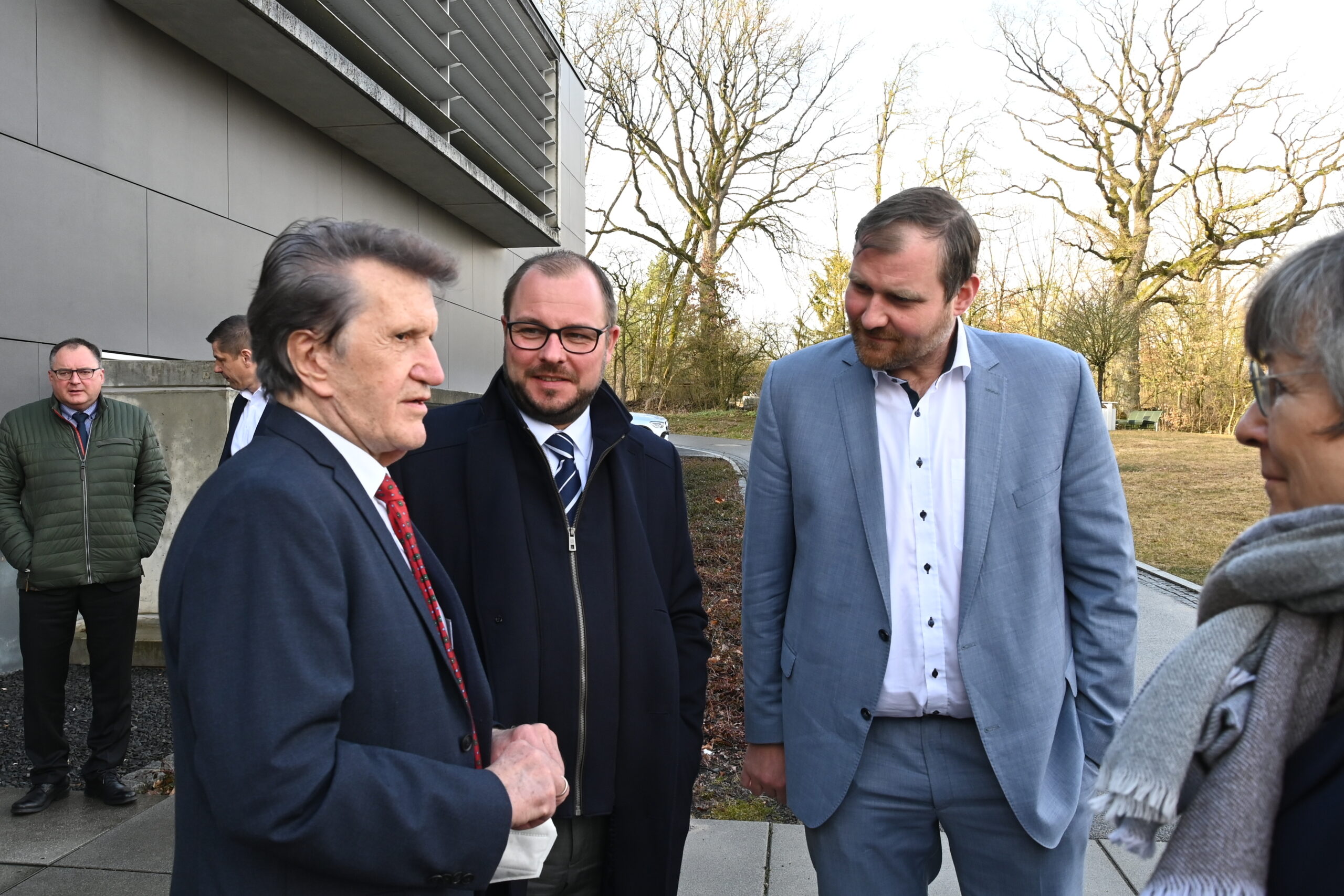 Eine Gruppe von Menschen in formeller Kleidung unterhält sich vor dem modernen Gebäude der Hochschule Coburg. Im Hintergrund sind Bäume und Gras zu sehen, was auf einen kalten oder frühen Frühlingstag schließen lässt. Die Personen scheinen in ein Gespräch vertieft zu sein.