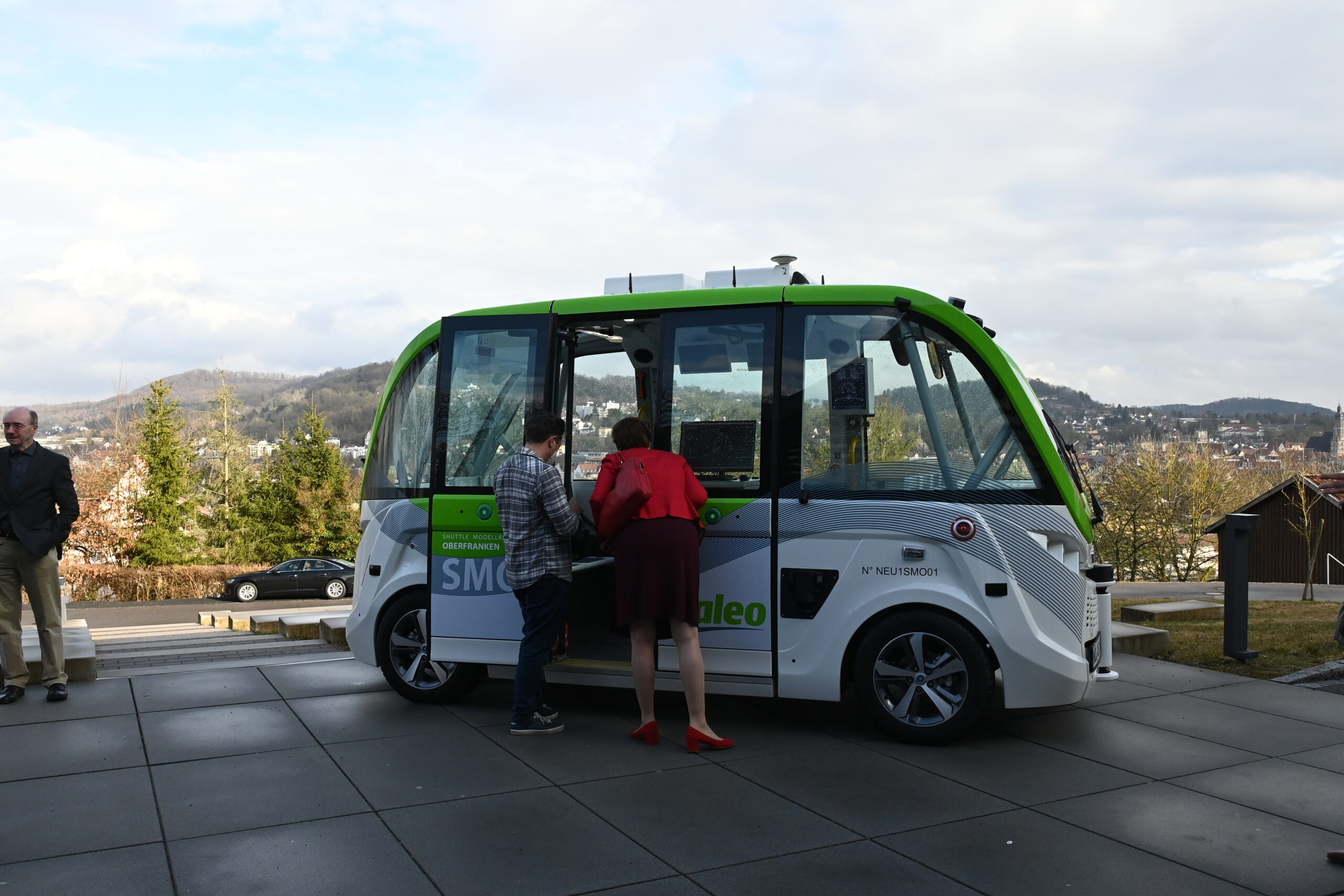 Zwei Personen untersuchen einen kleinen, fahrerlosen Shuttlebus im grün-weißen Design der Hochschule Coburg, der auf einer gepflasterten Fläche geparkt ist. Im Hintergrund ist eine malerische Aussicht auf Hügel und Bäume unter einem teilweise bewölkten Himmel zu sehen.