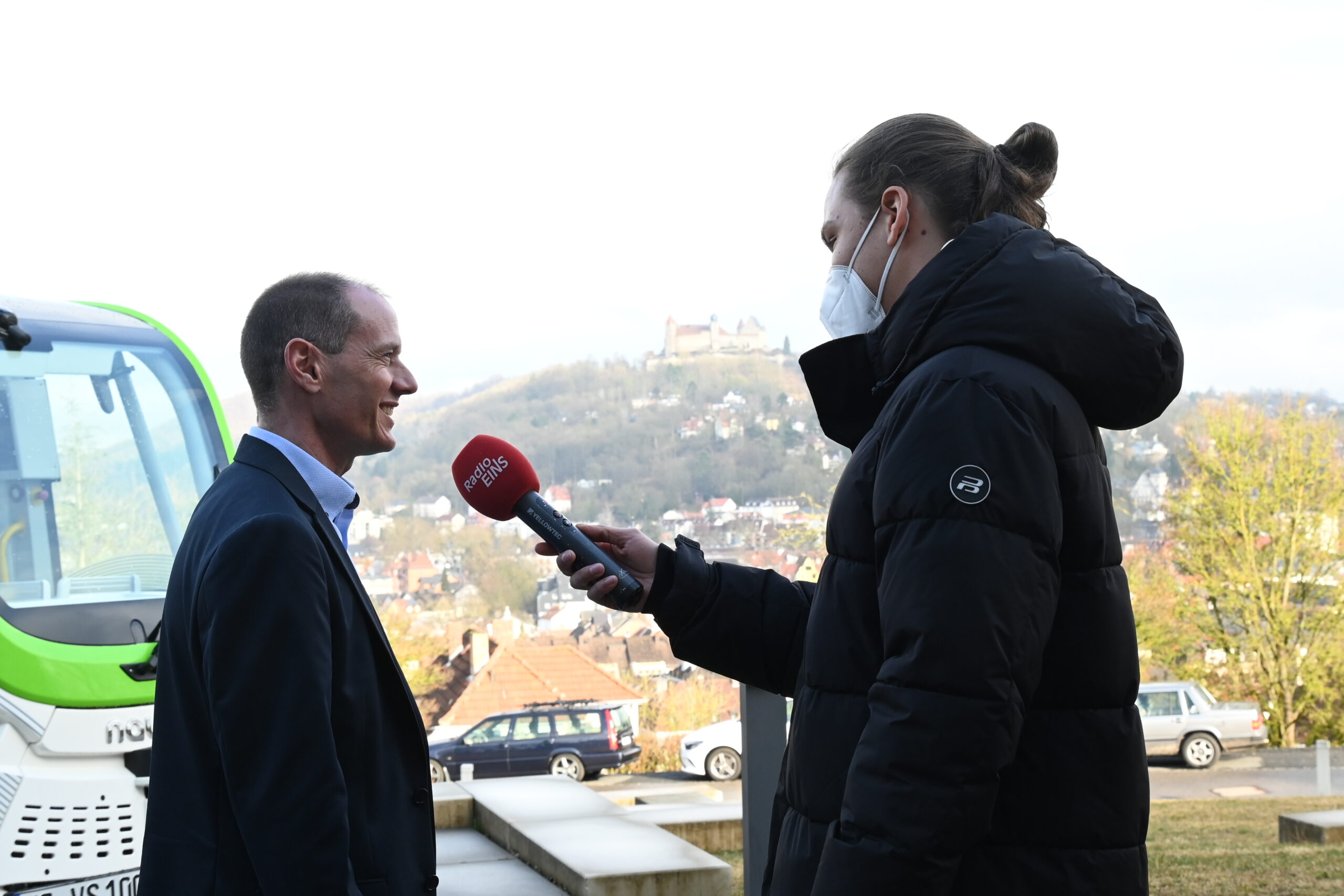 Ein Reporter in schwarzem Kittel und Gesichtsmaske, der ein Mikrofon mit der Aufschrift „ON AIR“ in der Hand hält, interviewt draußen einen Mann im Anzug. In der Nähe fährt ein grün-weißes Fahrzeug vorbei, die malerische Aussicht auf die Hochschule Coburg inmitten der Hügel bietet eine akademische Kulisse.