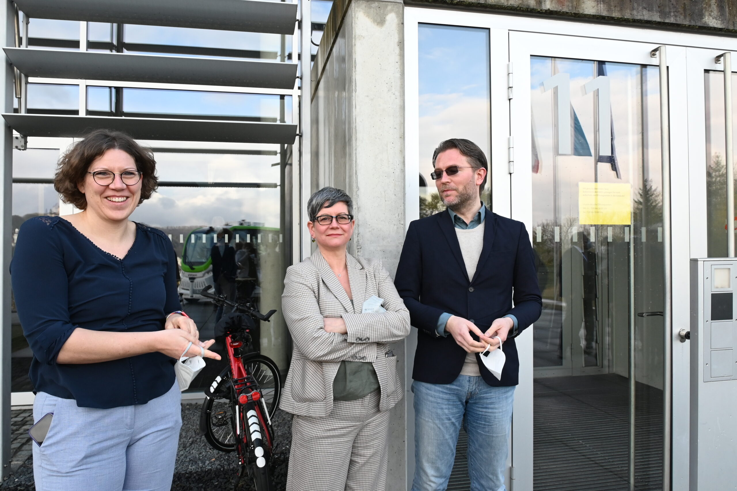 Drei Menschen stehen vor dem Eingang der Hochschule Coburg. Eine Frau links hält eine Maske, eine andere in der Mitte steht mit verschränkten Armen da. Ein Mann rechts trägt eine Sonnenbrille und strahlt kühles Selbstbewusstsein aus. In der Nähe lehnt ein Fahrrad an der Wand und vervollständigt diese alltägliche Szene.