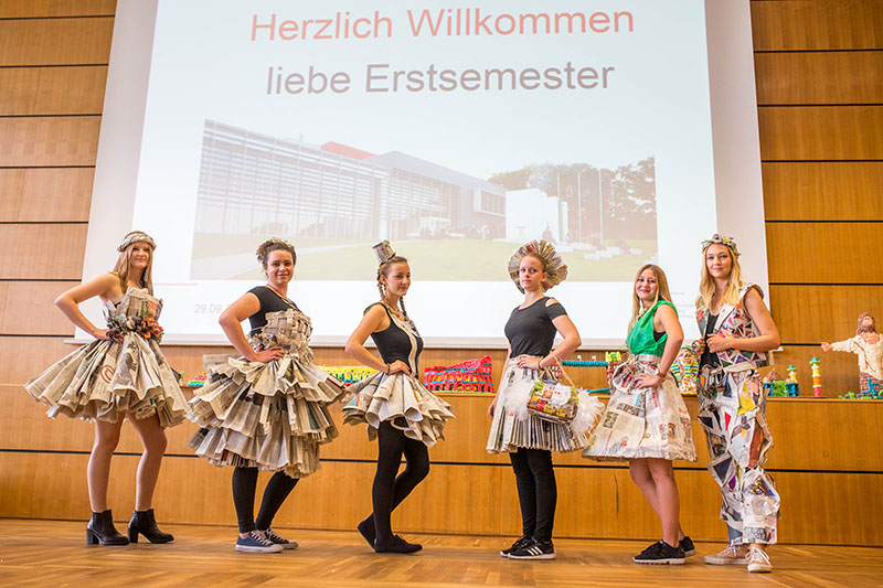 Auf der Bühne stehen sechs Personen in Kleidern aus Zeitungspapier. Hinter ihnen wird auf einem Bildschirm eine Begrüßungsnachricht auf Deutsch für die neuen Studierenden der Hochschule Coburg angezeigt. Die Kulisse scheint ein Auditorium oder ein Hörsaal zu sein, in dem die Menschen vor Erwartung und Aufregung über das bevorstehende akademische Leben summen.