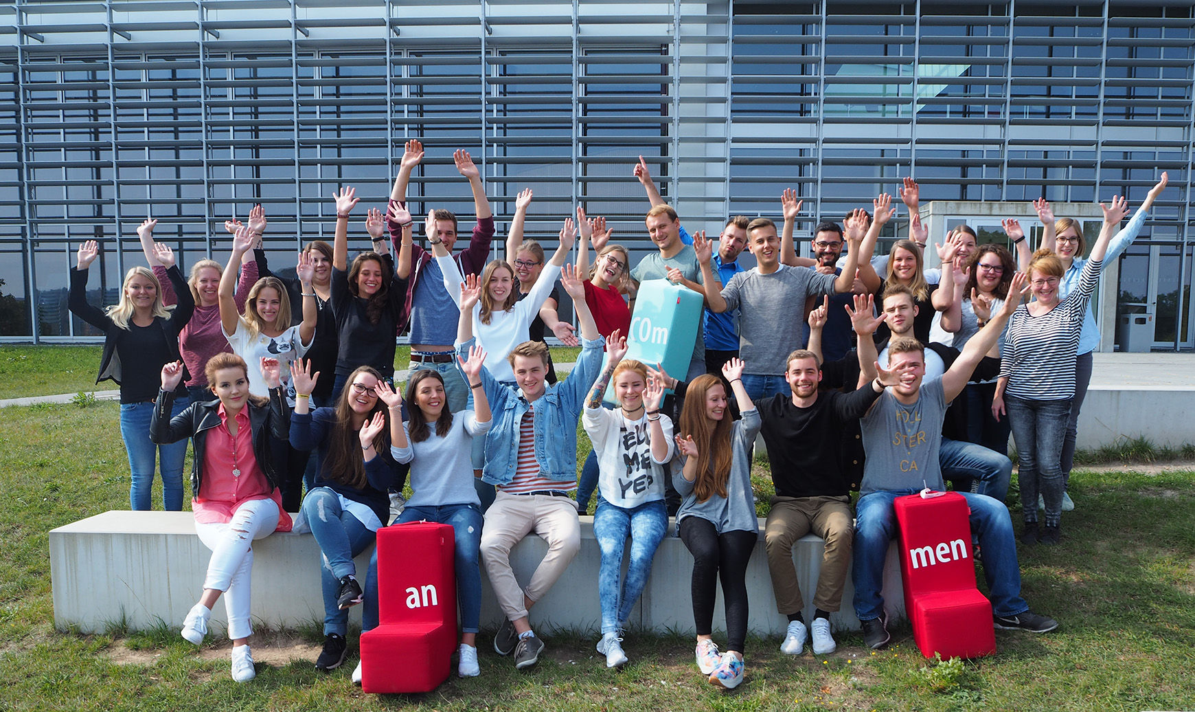 Vor dem modernen Gebäude mit den großen Fenstern der Hochschule Coburg hat sich eine Gruppe von Menschen im Freien versammelt. Sie lächeln und heben jubelnd die Hände, flankiert von zwei roten Blöcken mit weißen Buchstaben.