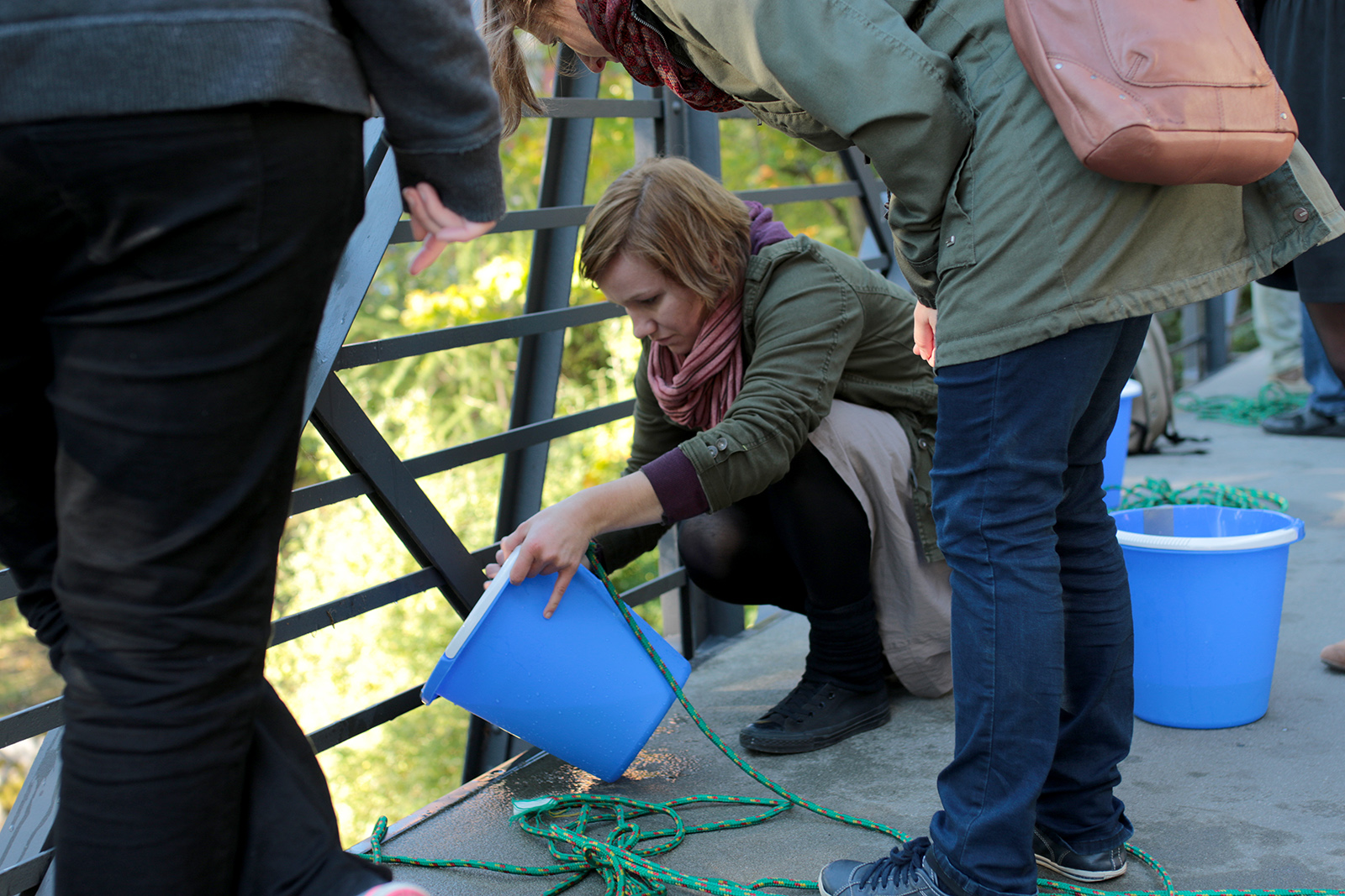 Eine Gruppe von Menschen der Hochschule Coburg steht auf einer Brücke und beschäftigt sich mit einer Aktivität mit einem blauen Eimer und grünen Seilen. Eine Person kniet, um den Eimer zu kippen, während andere herumstehen und mit der Aktivität beschäftigt sind. Im Hintergrund sind Baumblätter zu sehen.