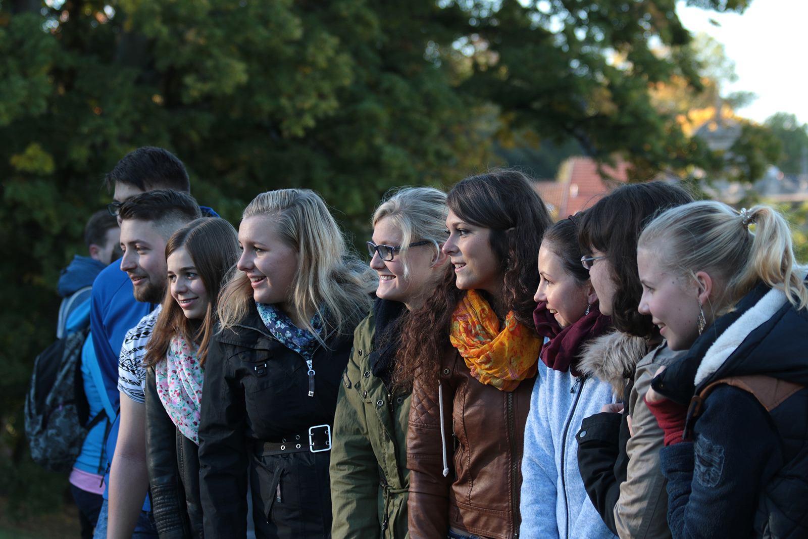 Eine Gruppe junger Erwachsener steht draußen in einer Reihe, lächelt und unterhält sich und verkörpert den lebendigen Geist der Hochschule Coburg. Sie sind in legere Herbstkleidung gekleidet, mit Bäumen und sanftem Abendlicht im Hintergrund.