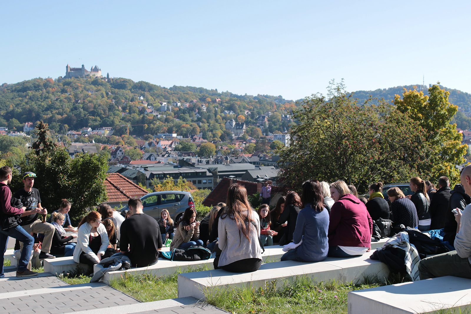 Eine Gruppe von Menschen sitzt auf Bänken in einem Park in der Nähe der Hochschule Coburg und genießt einen sonnigen Tag. Im Hintergrund thront ein Schloss in der Ferne auf einem Hügel, umgeben von einer malerischen Landschaft aus Bäumen und Dächern.