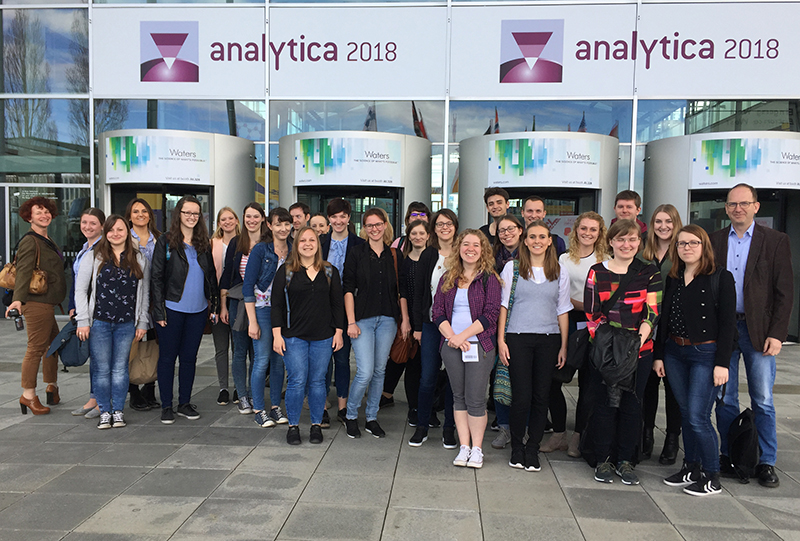 Eine Gruppe junger Erwachsener steht lächelnd vor dem Eingang zur Analytica 2018, einem Konferenzzentrum, über dessen Türen ein Schild prangt. Sie sind leger gekleidet und scheinen Studenten der Hochschule Coburg zu sein, die gespannt darauf sind, die neuesten Erkenntnisse der Branche zu erfahren.