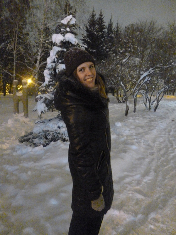 Eine Person in dunklem Mantel und Hut steht lächelnd in der Nähe des verschneiten Parks der Hochschule Coburg bei Nacht. Schneebedeckte Bäume und sanftes Licht sorgen für eine gemütliche Winteratmosphäre.