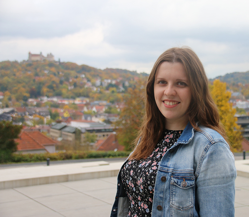 Eine Frau mit langen braunen Haaren lächelt in die Kamera. Sie trägt eine Jeansjacke über einem geblümten Oberteil. Sie steht vor einem malerischen Blick auf die Hochschule Coburg, mit einer Stadt am Hang und einem Schloss in der Ferne unter einem bewölkten Himmel.