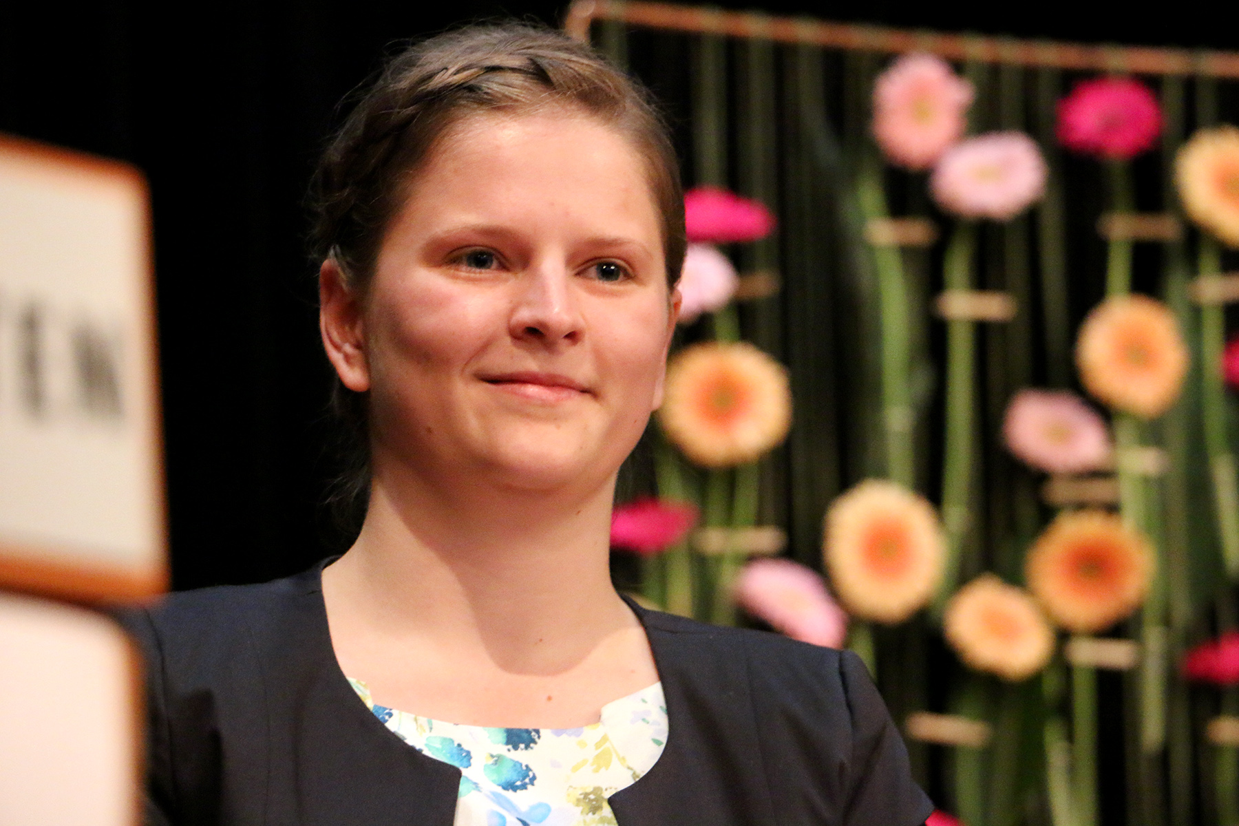 Eine Person mit kurzen Haaren steht vor einem dekorativen Hintergrund mit rosa und orangefarbenen Blumen, der an den lebhaften Campus der Hochschule Coburg erinnert, und lächelt leicht. Sie trägt eine dunkle Jacke über einem geblümten Oberteil.