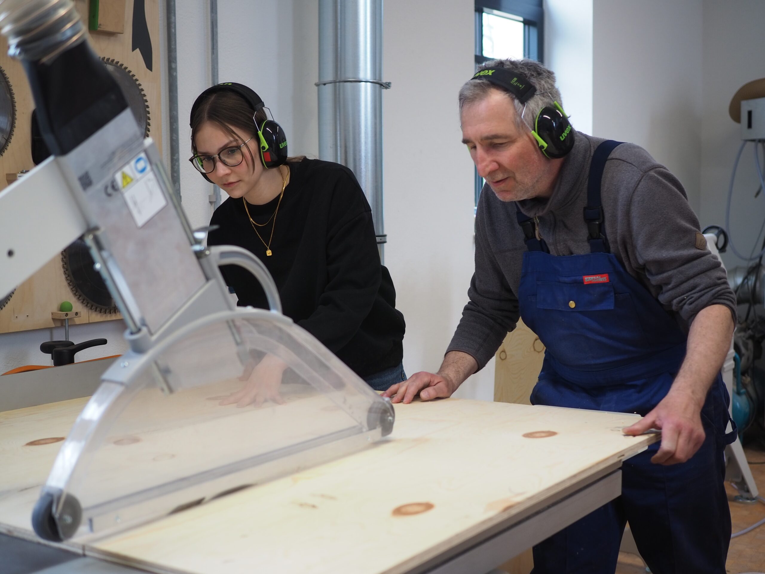 In einer Holzwerkstatt an der Hochschule Coburg tragen zwei Personen Ohrenschützer. Die jüngere Person ist konzentriert dabei, eine große Säge zu bedienen, um Sperrholz zu schneiden, während die ältere Person im blauen Overall die Arbeit mit Präzision überwacht. Sägeblätter und Werkzeuge füllen den Hintergrund dieser praktischen Lernumgebung.