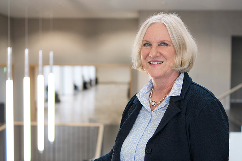 Eine ältere Frau mit kurzen weißen Haaren steht lächelnd in einem modernen, gut beleuchteten Innenraum der Hochschule Coburg. Sie trägt einen dunklen Blazer über einer hellblauen Bluse, was ihr einen professionellen Touch verleiht. Hängelampen und eine Treppe sorgen im Hintergrund für eine einladende Atmosphäre.