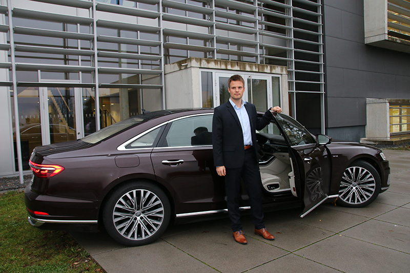 Ein Mann im Anzug steht neben der offenen Autotür einer Luxuslimousine, die auf einem gepflasterten Platz in der Nähe der Hochschule Coburg geparkt ist. Das Gebäude dahinter ist mit großen Fenstern und Metallpaneelen versehen. Das Auto ist dunkelbraun mit silbernen Felgen und strahlt vor der akademischen Kulisse Eleganz aus.