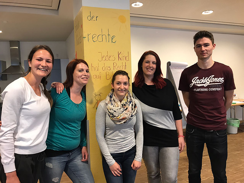 Eine Gruppe von fünf Personen, leger gekleidet, steht drinnen neben einer gelben Säule mit Schrift. Vier Frauen und ein Mann in einem kastanienbraunen T-Shirt lächeln herzlich. Die Umgebung erinnert an einen Unterrichts- oder Besprechungsraum der Hochschule Coburg.