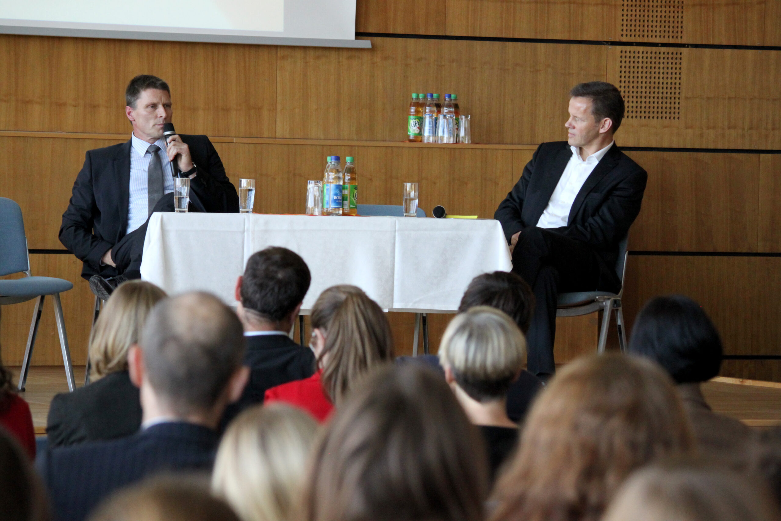 Zwei Männer in Anzügen sitzen auf einer Bühne der Hochschule Coburg und diskutieren. Einer spricht in ein Mikrofon, während der andere aufmerksam zuhört. Ein Tisch mit Wasserflaschen und Gläsern trennt sie, im Vordergrund sitzt ein gespanntes Publikum.