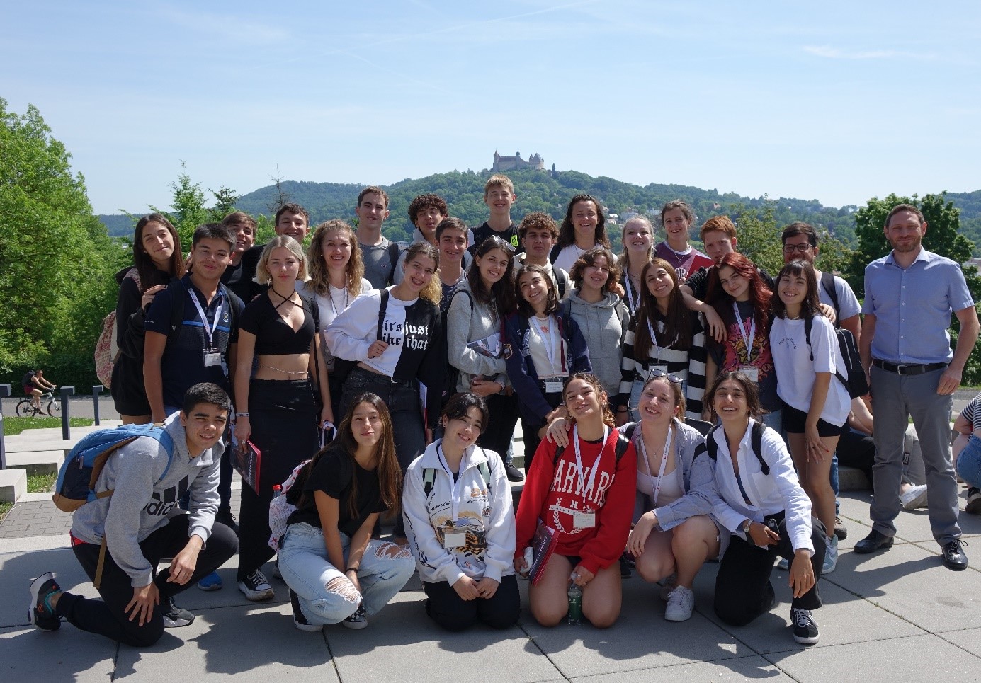 Eine Gruppe lächelnder Menschen, überwiegend Teenager, posiert gemeinsam im Freien vor einer Hügellandschaft mit einem Schloss im Hintergrund. In legerer Kleidung unter dem sonnigen Himmel verkörpern sie den lebendigen Geist, der oft an der Hochschule Coburg herrscht.