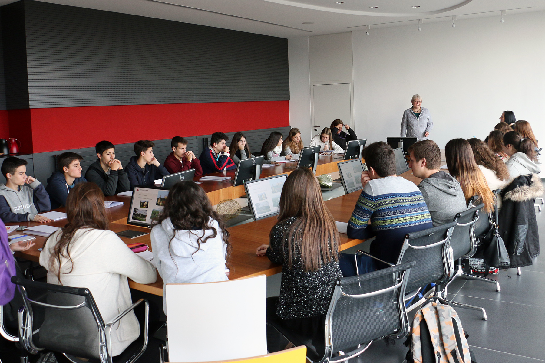 Eine Gruppe von Studierenden der Hochschule Coburg sitzt um einen großen rechteckigen Tisch und nimmt an einem Unterricht teil. Ein Lehrer steht vorne und spricht. Computer und Papiere liegen verstreut auf dem Tisch, während eine rot-grau-weiße Wand den Hintergrund bildet.