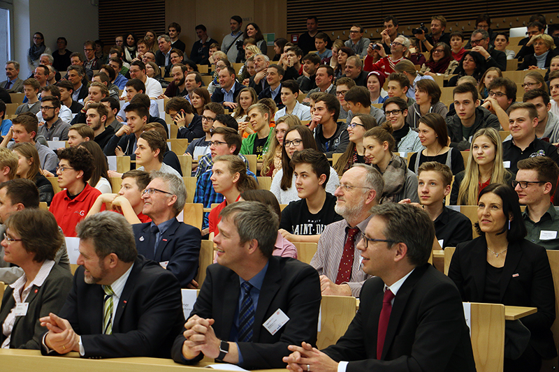 Ein Hörsaal der Hochschule Coburg ist mit einem großen Publikum gefüllt, darunter sowohl Studierende als auch Fachleute. Viele sitzen in Sitzreihen, blicken aufmerksam nach vorne, einige lächeln, andere konzentrieren sich auf eine Präsentation.