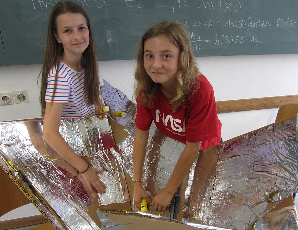 In einem Hörsaal der Hochschule Coburg arbeiten zwei Mädchen gemeinsam an einem Projekt mit glänzender Folie. Eine trägt ein gestreiftes Shirt, die andere ist rot. Im Hintergrund ragt eine Tafel mit Berechnungen hervor, während sie mit einem leichten Lächeln von ihrer Arbeit aufblicken.