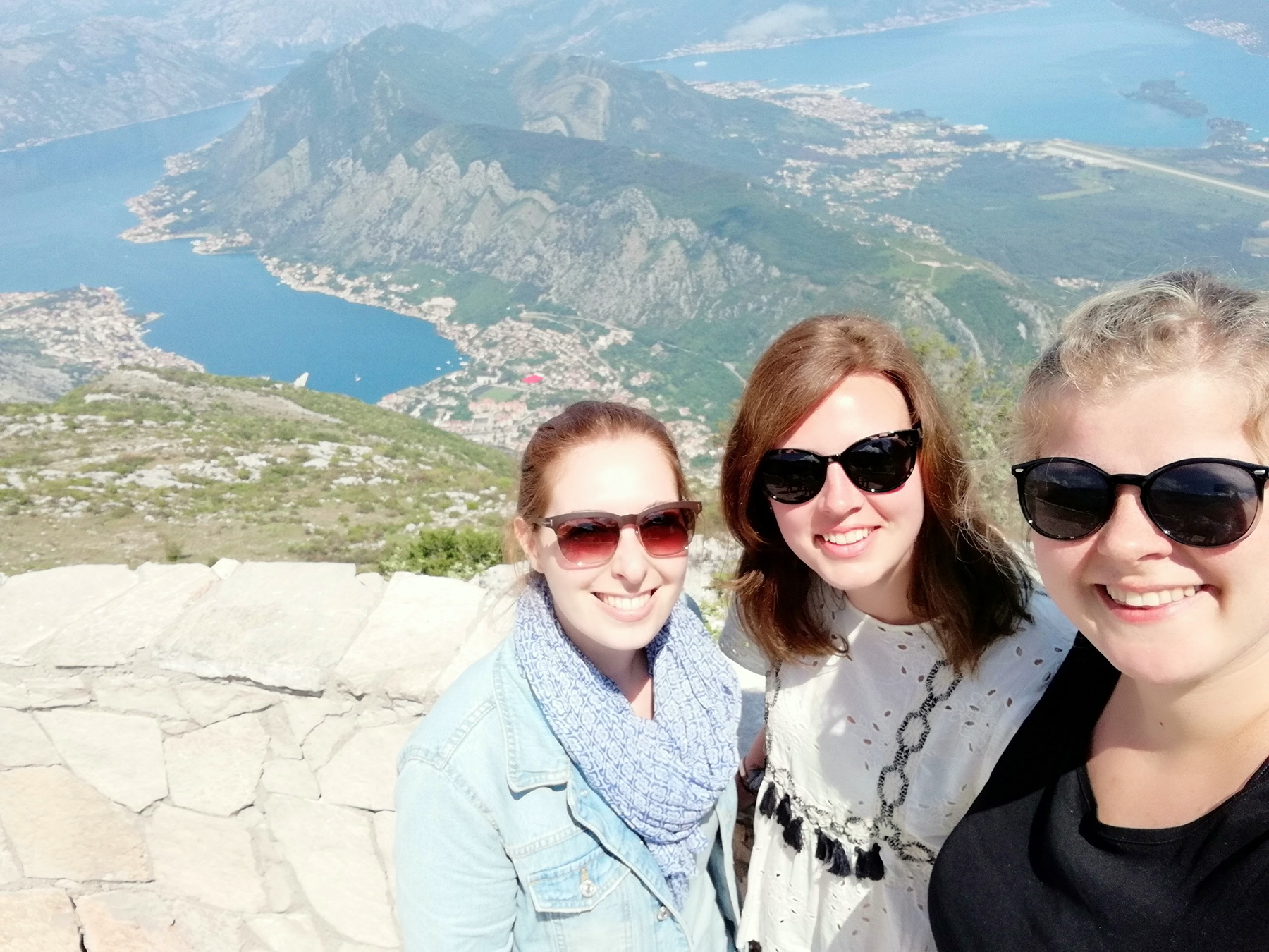 Drei Frauen mit Sonnenbrillen machen ein Selfie auf einem Berg. Dahinter ist der malerische Blick auf die Hochschule Coburg mit ihrer üppigen grünen Umgebung und dem ruhigen See zu sehen. Der sonnige Tag unterstreicht den klaren blauen Himmel und die Lebendigkeit dieser malerischen Landschaft.