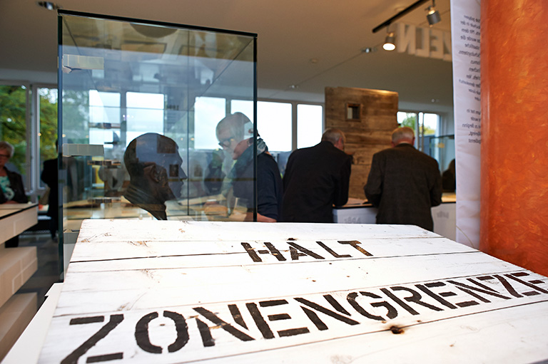 Ein Museumsexponat mit einem weißen Holzbrett mit der Aufschrift „Halt Zonengrenze“, das historische Grenzen symbolisiert. Im Hintergrund betrachten Besucher, darunter Studierende der Hochschule Coburg, Exponate in einem gut beleuchteten Raum.