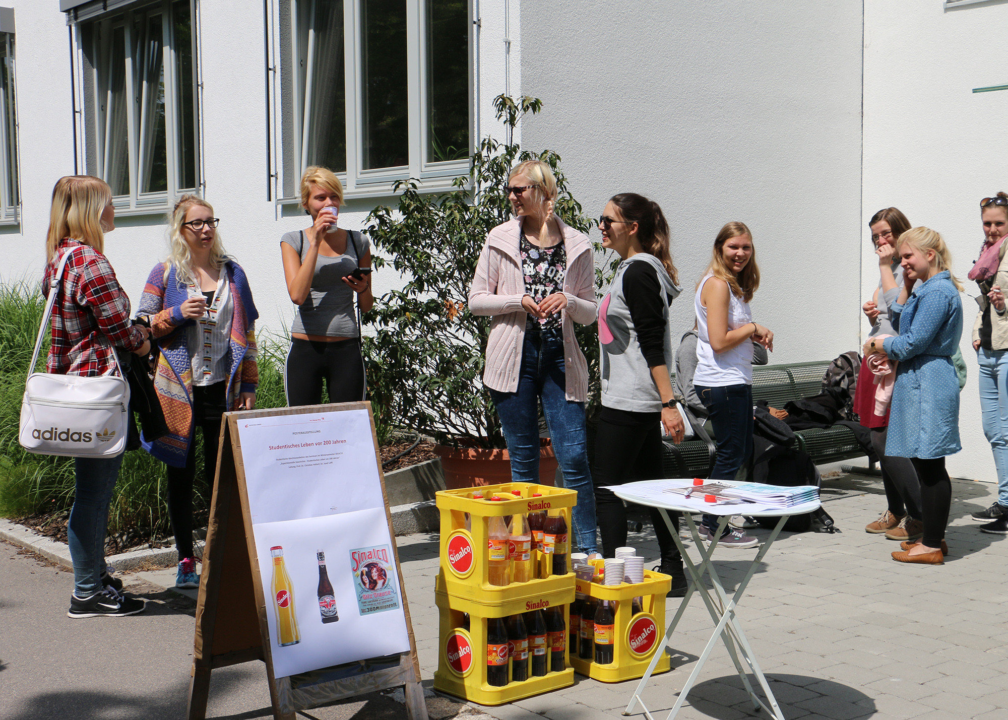 Vor einem Gebäude in der Nähe der Hochschule Coburg trifft sich eine Gruppe von Menschen zu einem lockeren Beisammensein. Getränkekisten und ein Werbeschild sind ausgestellt, auf einem kleinen Tisch liegen weitere Gegenstände. Mehrere Teilnehmer unterhalten sich angeregt.