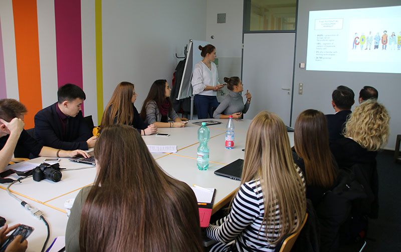 In einem modernen Raum mit bunt gestreiften Wänden sitzt eine Gruppe von Menschen um einen Konferenztisch. An der Hochschule Coburg steht eine Person ganz vorne und präsentiert Folien auf einer Leinwand. Die Teilnehmer machen sich Notizen und hören aufmerksam zu.