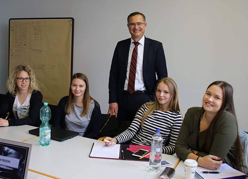 Ein Mann im Anzug lächelt hinter einem Tisch, an dem vier strahlende junge Frauen in einem Besprechungsraum der Hochschule Coburg sitzen. Laptops, Notizbücher und Wasserflaschen liegen verstreut auf dem Tisch, im Hintergrund steht ein Flipchart voller Notizen.