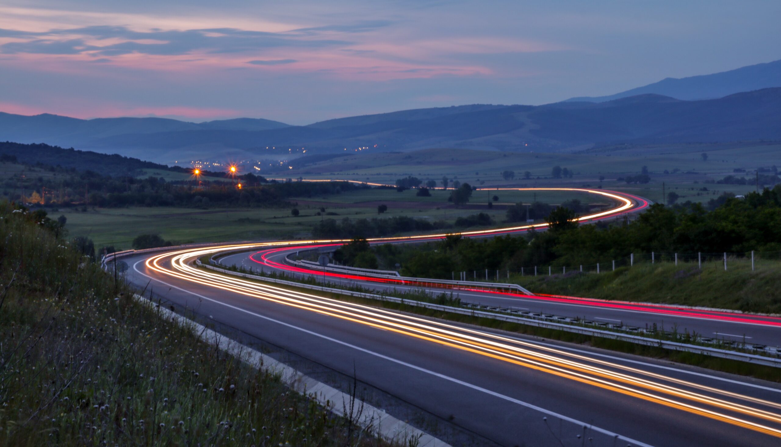 Eine Langzeitbelichtung einer Autobahn in der Abenddämmerung in der Nähe der Hochschule Coburg, bei der die Autolichter dynamische rote und weiße Streifen erzeugen. Die Straße schlängelt sich durch eine ländliche Landschaft mit sanften Hügeln im Hintergrund unter einem farbenfrohen Abendhimmel.