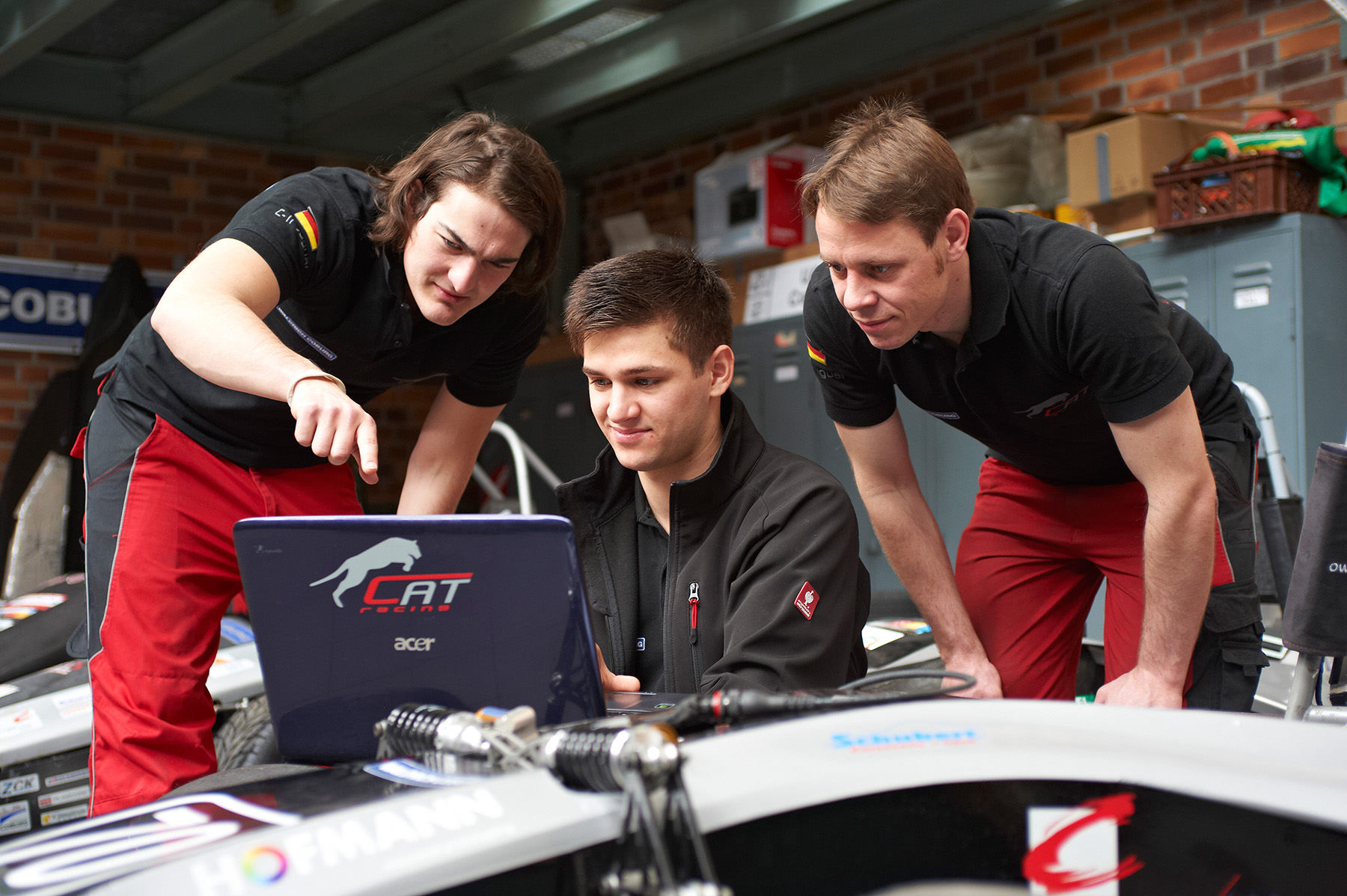 In einer Garage sitzen drei Leute um einen Laptop mit dem „CAT“-Logo auf einem Rennwagen herum. Sie arbeiten eng zusammen und analysieren Daten. Die Kulisse aus Backsteinmauern und Lagerschränken weckt einen akademischen Geist, der an das Innovationszentrum der Hochschule Coburg erinnert.