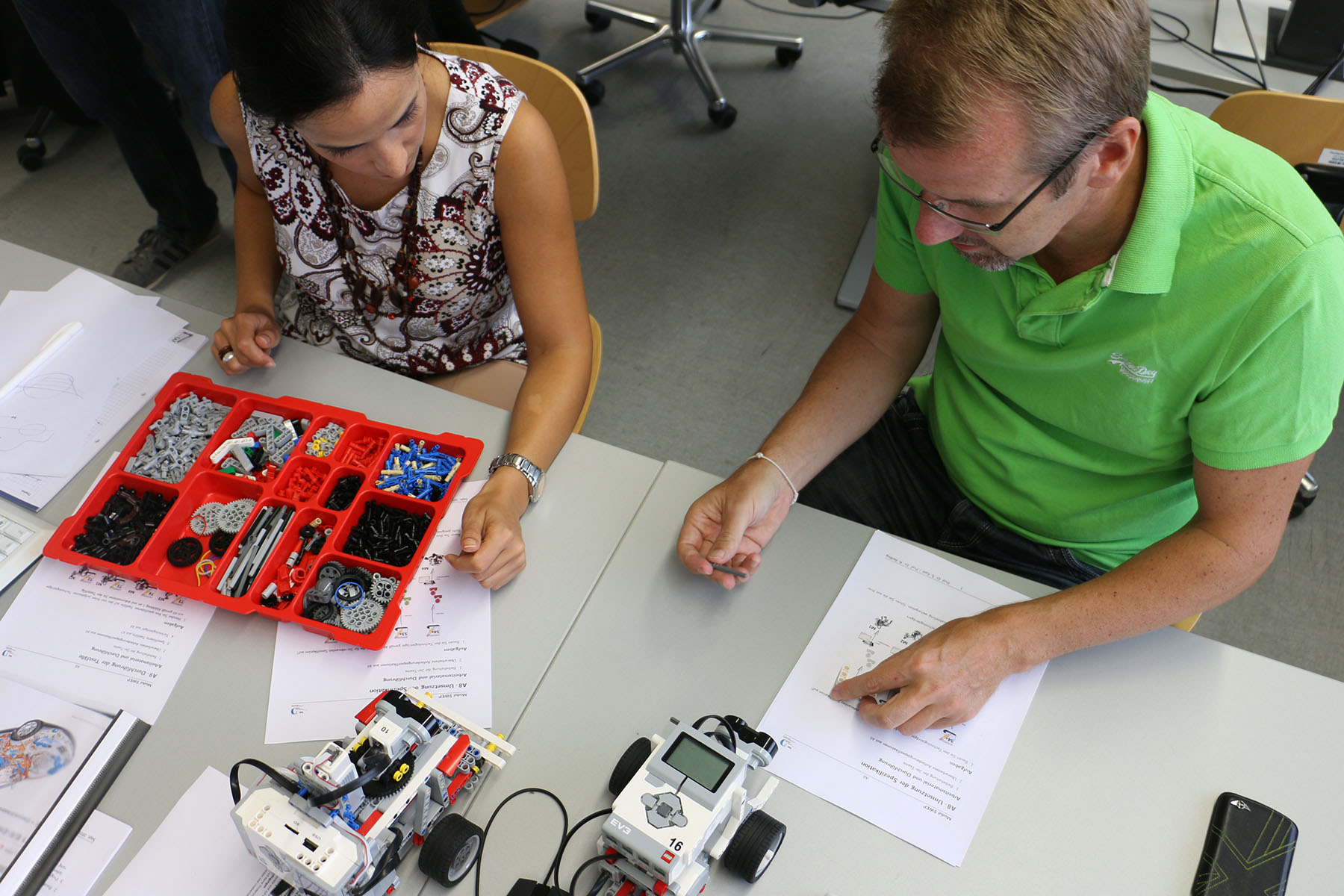 Two people sit at a table at Hochschule Coburg, immersed in their robotics kits. They sort through various components in a red organizer tray and build small robots, with instruction papers spread across the table.