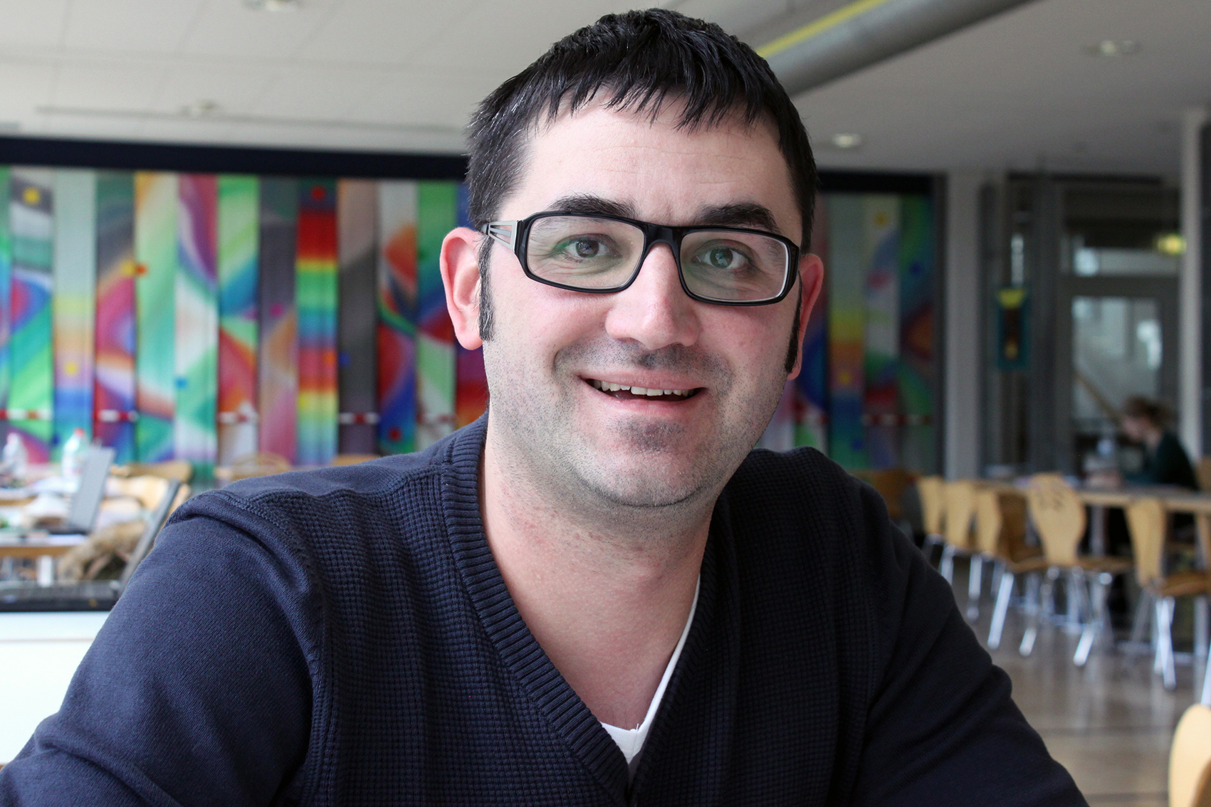 Ein Mann mit kurzen dunklen Haaren, Brille und dunklem Pullover sitzt lächelnd in einem hellen Raum mit abstrakter Kunst an der Wand. Mit Tischen und Stühlen im Hintergrund erinnert die Umgebung an einen lebendigen Raum der Hochschule Coburg.