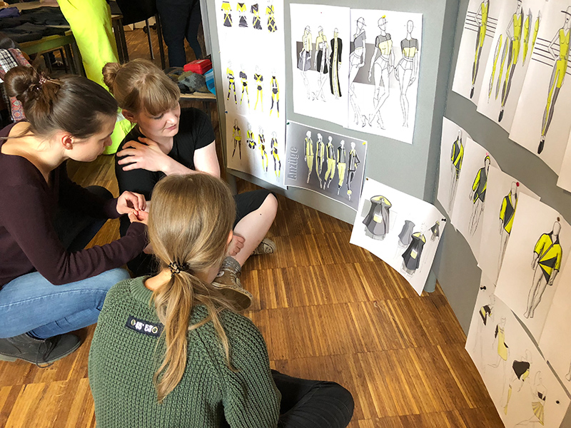 Drei Personen sitzen auf dem Holzboden eines Studios der Hochschule Coburg und diskutieren über Modeskizzen, die an der Wand hängen. Die Entwürfe zeigen Kleidung in Gelb- und Schwarztönen und schaffen eine kollaborative Atmosphäre, während sie gemeinsam kreative Ideen entwickeln.