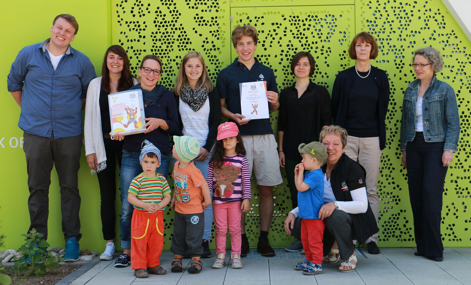 Eine Gruppe Erwachsener, manche mit Zertifikaten in den Händen, steht stolz hinter ein paar bunt gekleideten Kindern. Sie posieren vor einer grünen, perforierten Wand an der Hochschule Coburg.