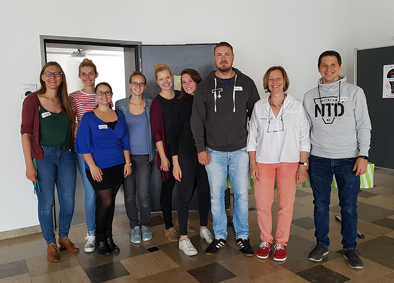 Eine Gruppe von neun Personen der Hochschule Coburg steht drinnen und lächelt in die Kamera. Sie sind leger gekleidet und stehen vor einer schlichten Wand. Einige halten kleine Taschen in den Händen. Der Boden unter ihnen ist mit einem gemischten Fliesenmuster versehen, was dem Ambiente einen Hauch von Charme verleiht.