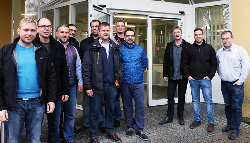 Vor dem Glastüreingang der Hochschule Coburg steht eine Gruppe von zehn Männern. Sie sind leger gekleidet, die meisten tragen Sakko. Im Hintergrund ist eine gelbe Wand zu sehen, die einen Blick ins Innere des Gebäudes bietet.