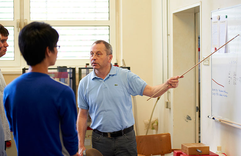 Ein Mann im hellblauen Poloshirt steht in einem Hörsaal der Hochschule Coburg. Er hält einen Zeigestab in der Hand und erklärt etwas auf einer Tafel voller Diagramme. Zwei Studierende sitzen ihm gegenüber und hören aufmerksam zu.