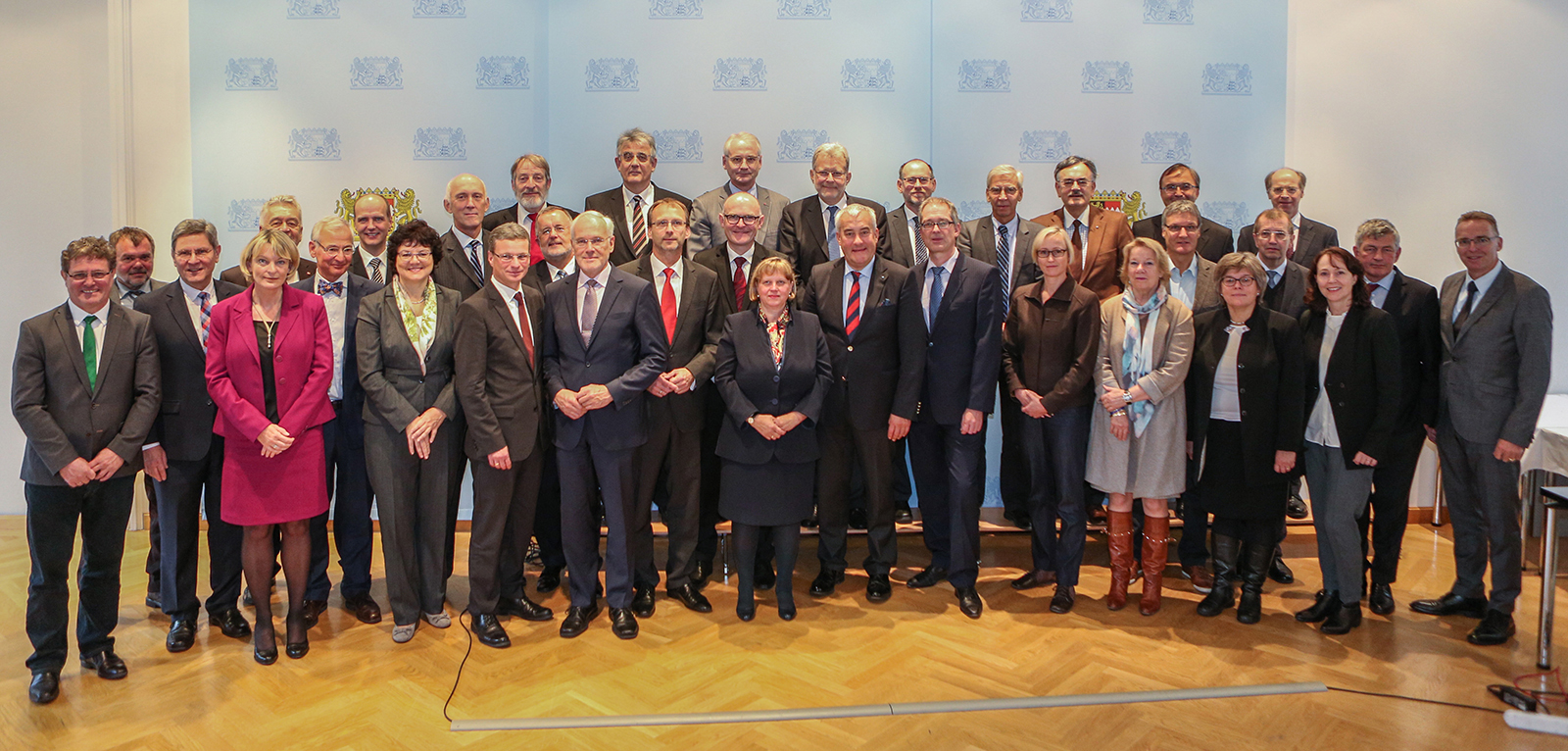 Eine Gruppe formell gekleideter Personen steht zusammen und posiert für ein Foto in einem gut beleuchteten Raum mit Parkettboden und einem Hintergrund mit dem Logomuster der Hochschule Coburg.