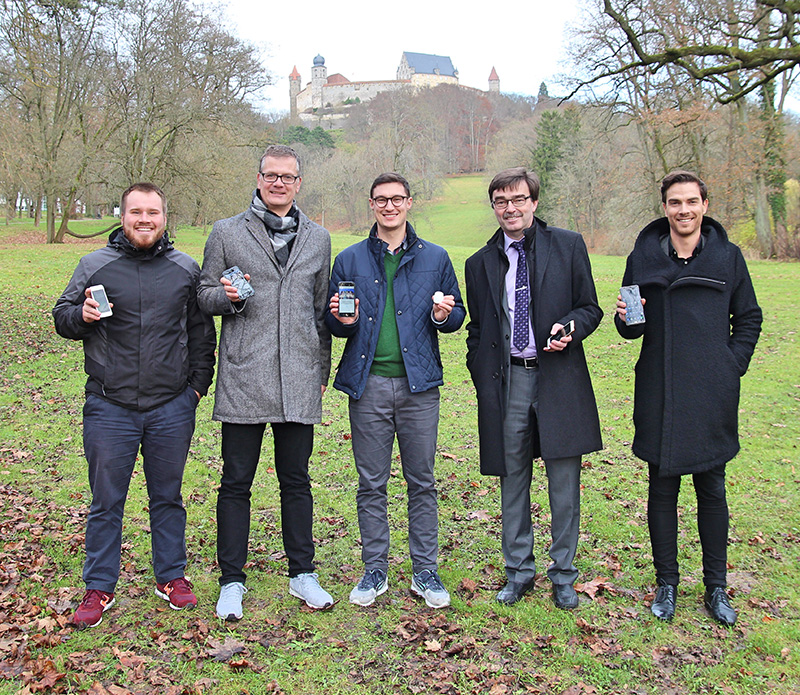 Fünf Männer stehen in einem Park und halten kleine Geräte in der Hand. Sie sind in Wintermäntel und Freizeit- oder Businesskleidung gekleidet. Hinter ihnen liegt das historische Gebäude der Hochschule Coburg auf einem Hügel, umgeben von kahlen Bäumen. Der Boden ist ein Teppich aus verstreuten Blättern.