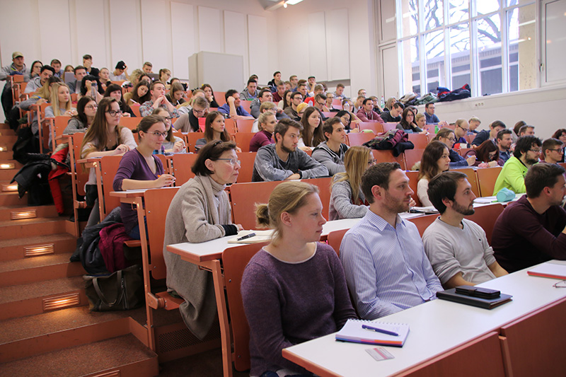 Im großen Hörsaal der Hochschule Coburg herrscht reges Treiben, denn die Studierenden hören aufmerksam zu und machen sich Notizen. Die Sitzreihen sind abgestuft, die meisten haben Notizbücher oder Laptops aufgeklappt, während durch die großen Fenster auf einer Seite Tageslicht hereinströmt.