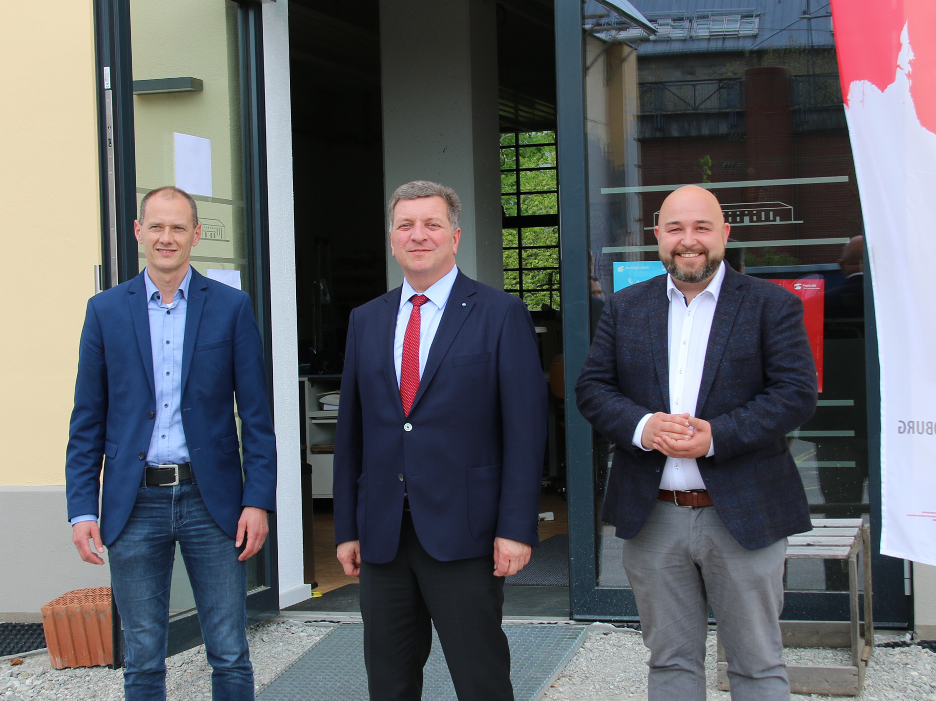 Drei Männer in Anzügen stehen lächelnd vor einem Gebäude mit einer Glastür. Der Mann links trägt Jeans und ein blaues Hemd, der Mann in der Mitte mit einer roten Krawatte hält Materialien der Hochschule Coburg in der Hand und der Mann rechts trägt ein weißes Hemd ohne Krawatte. Ein rot-weißes Schild ist teilweise sichtbar.