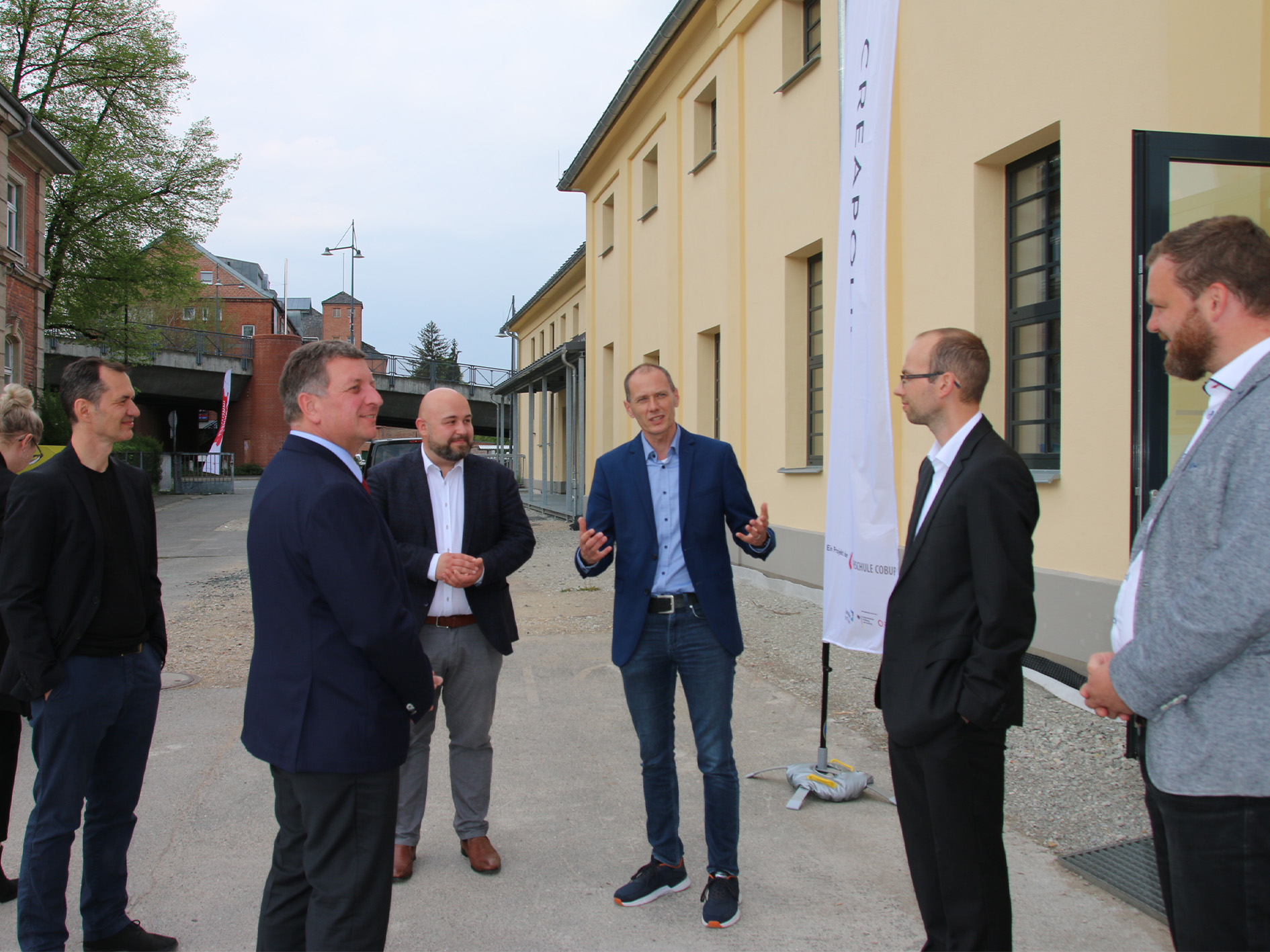 Eine Gruppe von sechs Männern in Geschäftskleidung steht im Freien in der Nähe eines gelben Gebäudes und unterhält sich, wahrscheinlich über Themen im Zusammenhang mit der Hochschule Coburg. Sie scheinen sich in einem Hofbereich zu befinden, in dem ein weißes Banner zu sehen ist. Im Hintergrund sind Bäume und ein weiteres Backsteingebäude zu sehen.