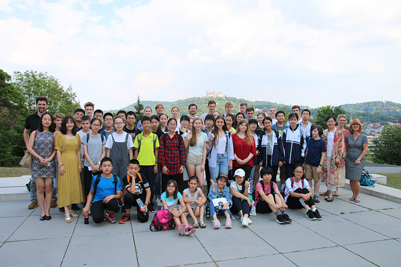 Eine große Gruppe von Studierenden und Lehrkräften der Hochschule Coburg posiert im Freien auf einer gepflasterten Fläche, im Hintergrund eine malerische Landschaft aus Bäumen und Hügeln. Der Himmel ist teilweise bewölkt. Die bunt gemischte Gruppe scheint auf einem Schulausflug oder einer Exkursion zu sein.