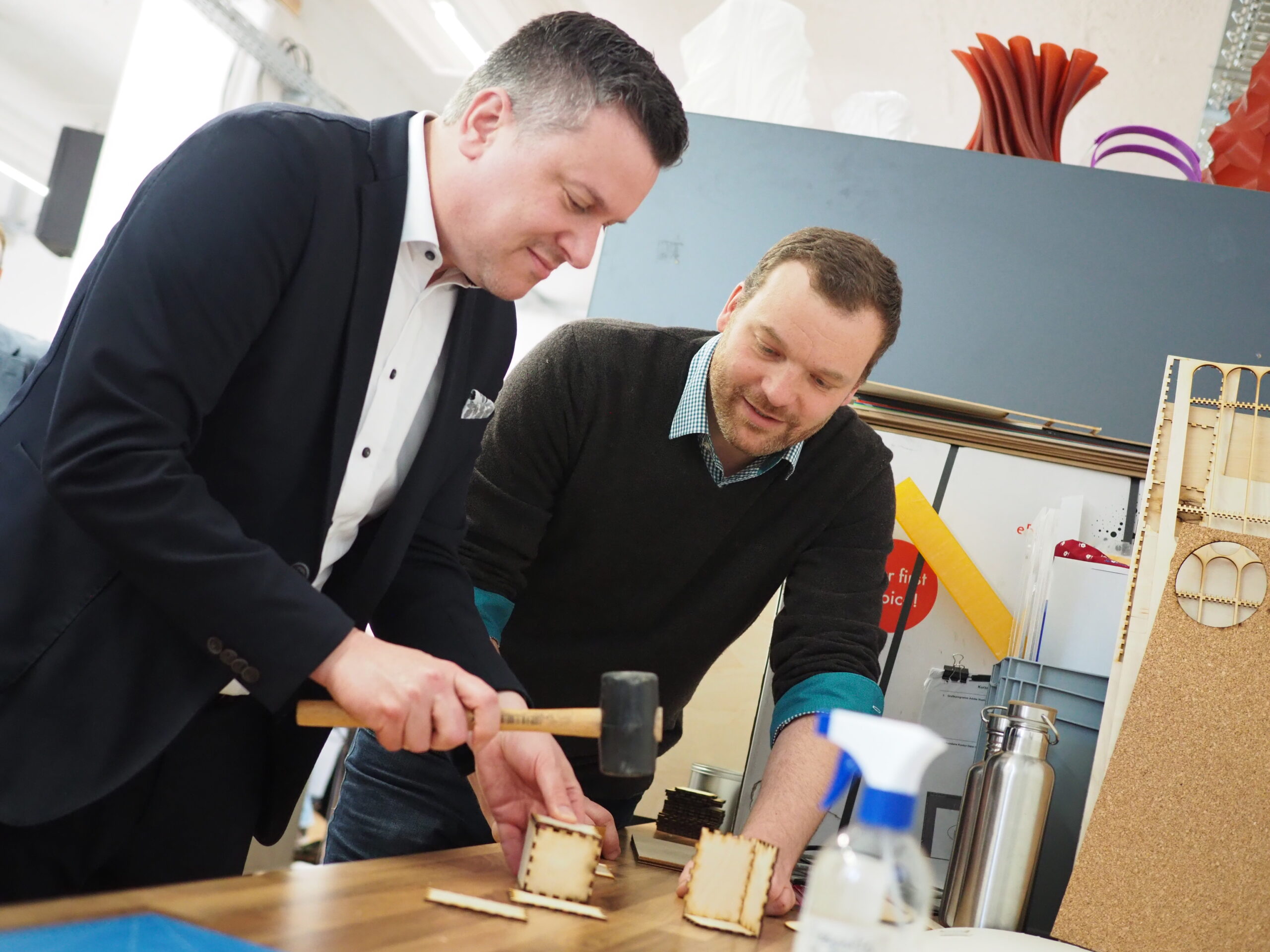 Zwei Männer bauen Holzmodelle auf einem Tisch zusammen und verkörpern damit die sorgfältige Handwerkskunst, die an der Hochschule Coburg gelehrt wird. Einer der Männer benutzt einen Hammer, während der andere aufmerksam zusieht. Der Tisch ist mit Werkzeugen und Materialien übersät, und ein Regal mit bunten Objekten bildet den Hintergrund ihrer konzentrierten Arbeit.