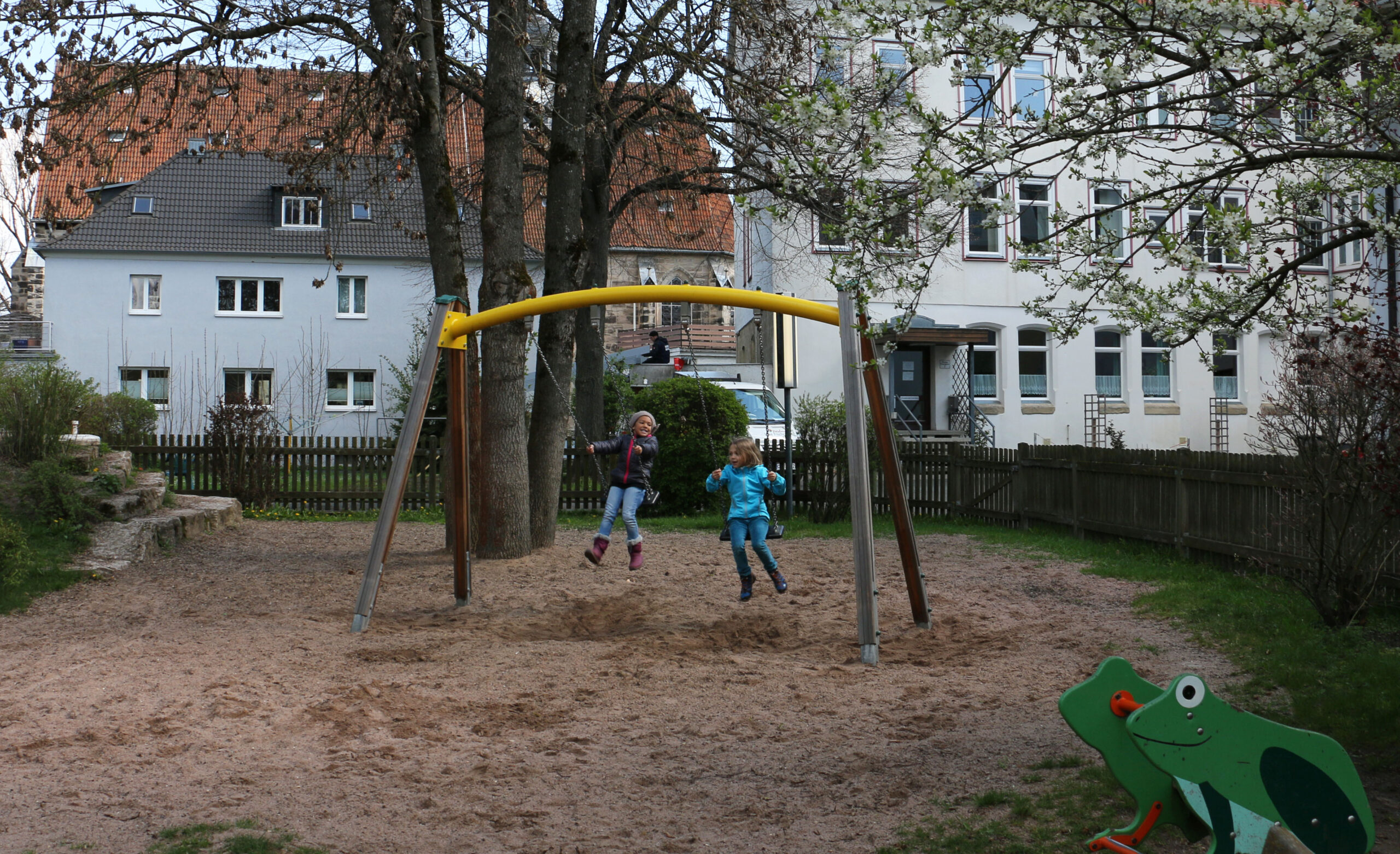 Zwei Kinder schaukeln fröhlich auf einer gelben Schaukel auf einem von Bäumen und Gebäuden umgebenen Spielplatz in der Nähe der Hochschule Coburg. Der Himmel ist klar und der Boden ist mit Sand bedeckt. Ein froschförmiges Spielgerät verleiht der Szene im Vordergrund Charme.