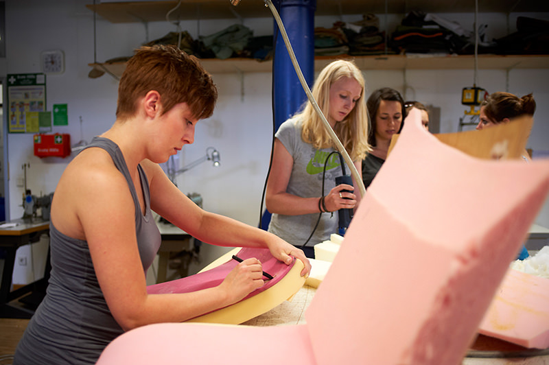 Eine Gruppe von Frauen an der Hochschule Coburg arbeitet fleißig in einer Werkstatt und fertigt einen rosa Schaumstoffstuhl. Eine Frau malt auf dem Schaumstoff, während andere konzentriert im Hintergrund stehen, umgeben von Regalen voller Werkzeuge und Materialien.