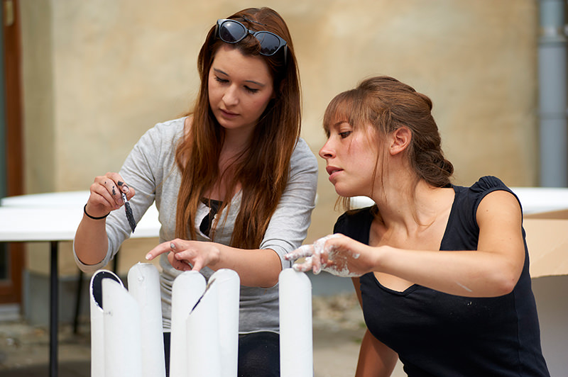 Zwei Frauen sind in ein künstlerisches Projekt im Freien vertieft, das vielleicht von ihrer Zeit an der Hochschule Coburg inspiriert wurde. Eine hält einen Pinsel, während die andere mit ihren Fingern Farbe auf vertikale zylindrische Formen auf einem Tisch aufträgt. Beide sind völlig in ihr kreatives Unterfangen vertieft.