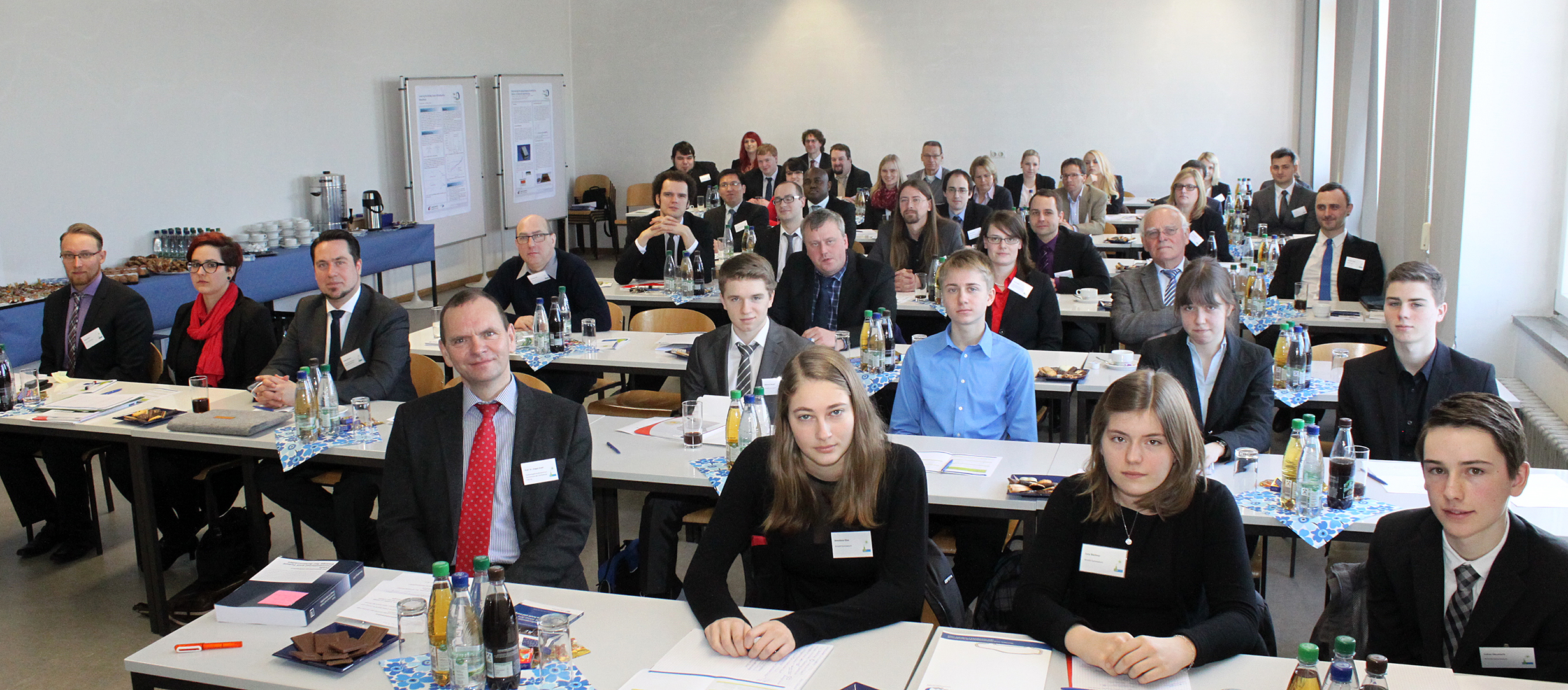 Eine gemischte Gruppe von Menschen sitzt in einem Konferenzraum der Hochschule Coburg und blickt in die Kamera. Die Tische sind mit Unterlagen, Getränken und Snacks ausgestattet. Im Hintergrund sind auf Beistelltischen Poster und Erfrischungen aufgestellt. Die Atmosphäre ist professionell und formell.